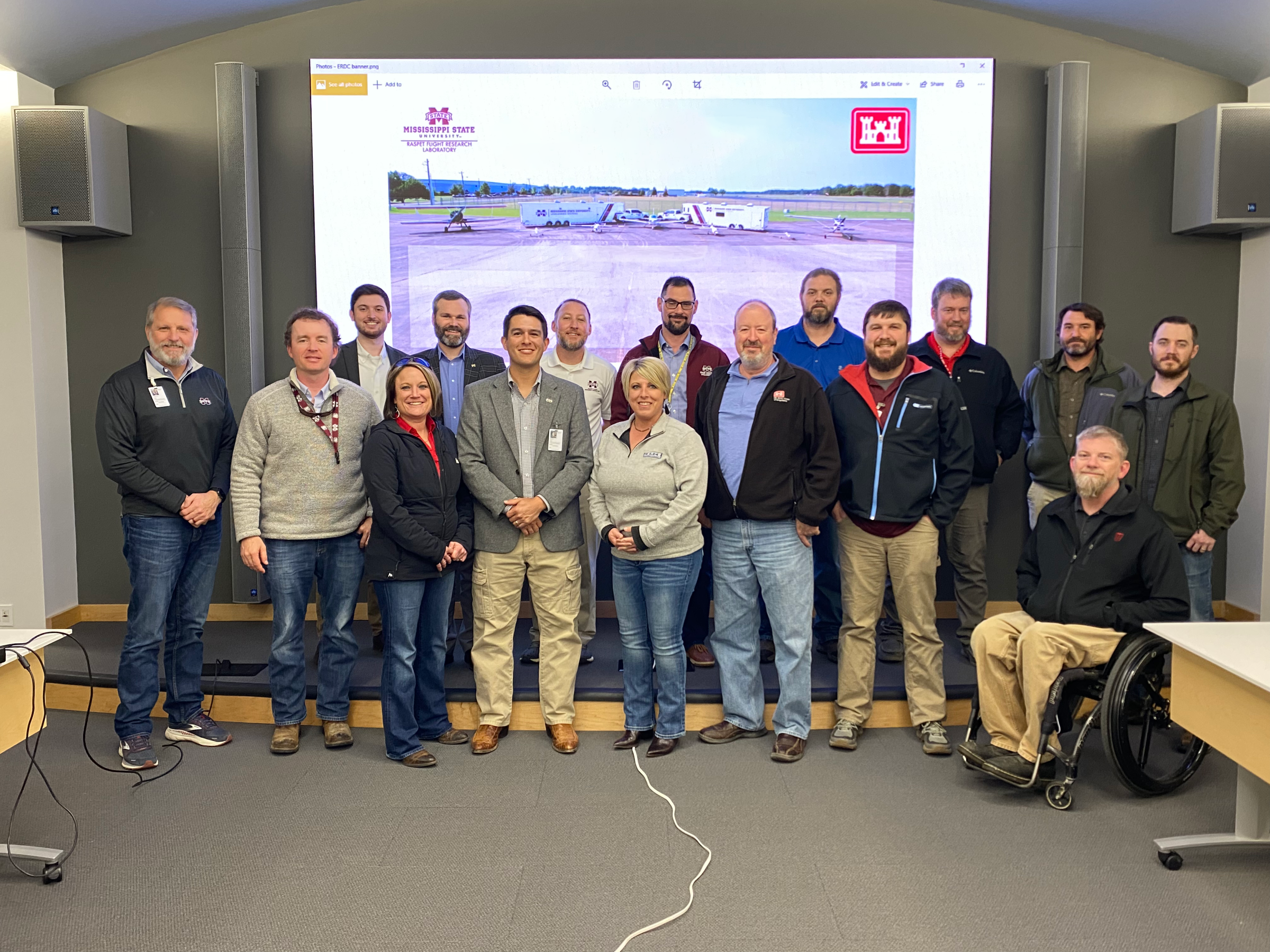 A group of 16 people posed in front of a projection screen displaying a graphic containing Raspet Flight Lab and Engineer Research and Development Center logos. 