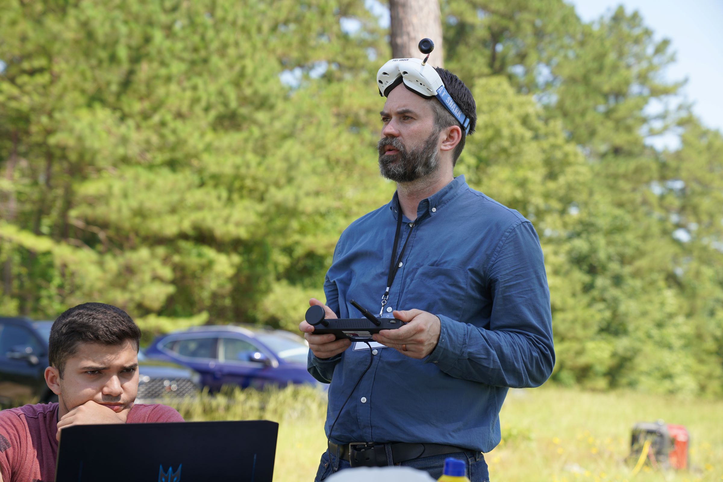 A drone operator holds a controller while a teammate seated beside him looks at a laptop. 