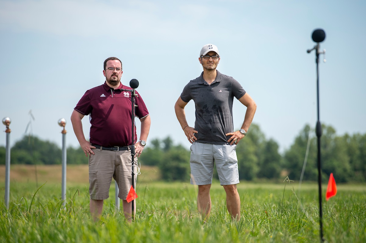 Two men stand in a field with an acoustic microphone array. 